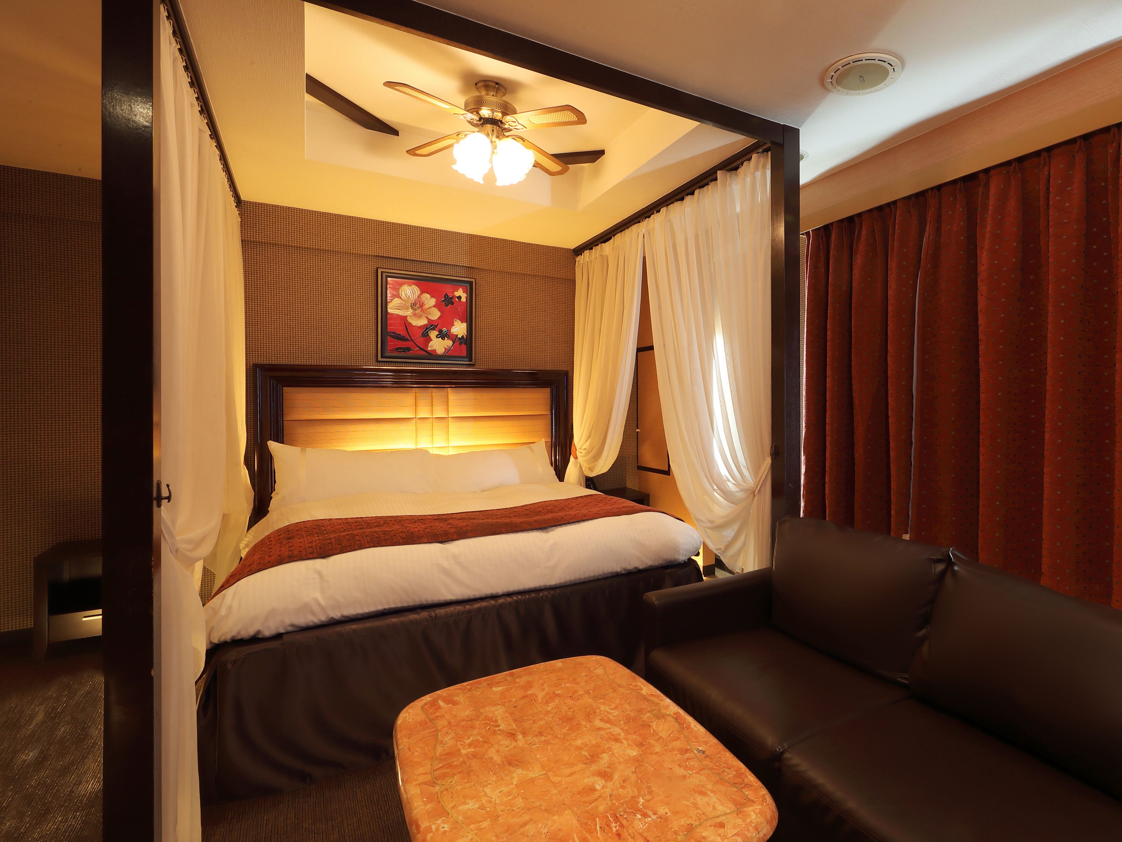 Une chambre d’hôtel avec un lit, un ventilateur de plafond, un tableau et un canapé en cuir marron.
