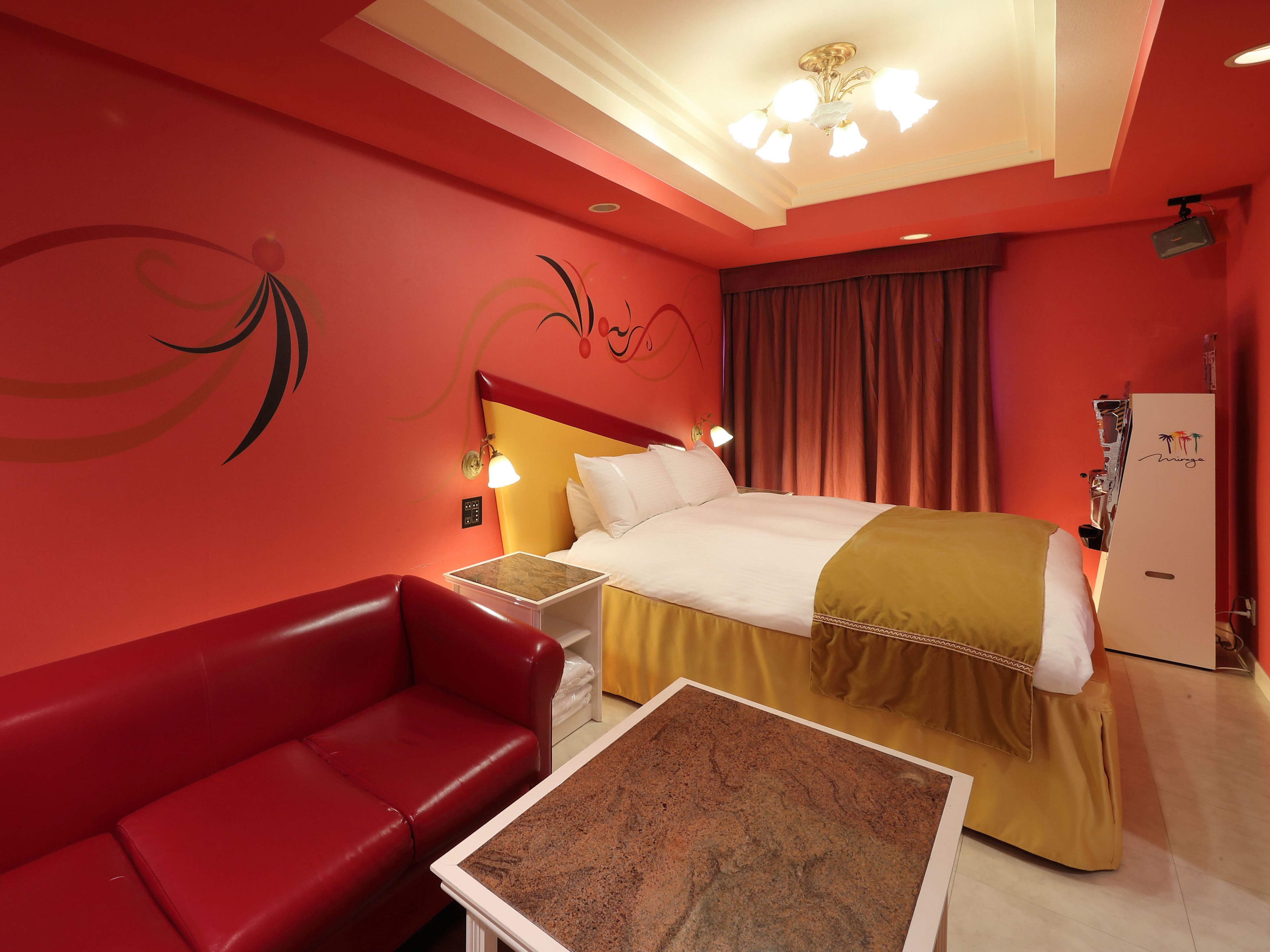 A hotel room with a red wall, a bed with a white and yellow bedspread, a red leather sofa, a small table, and a ceiling light fixture.
