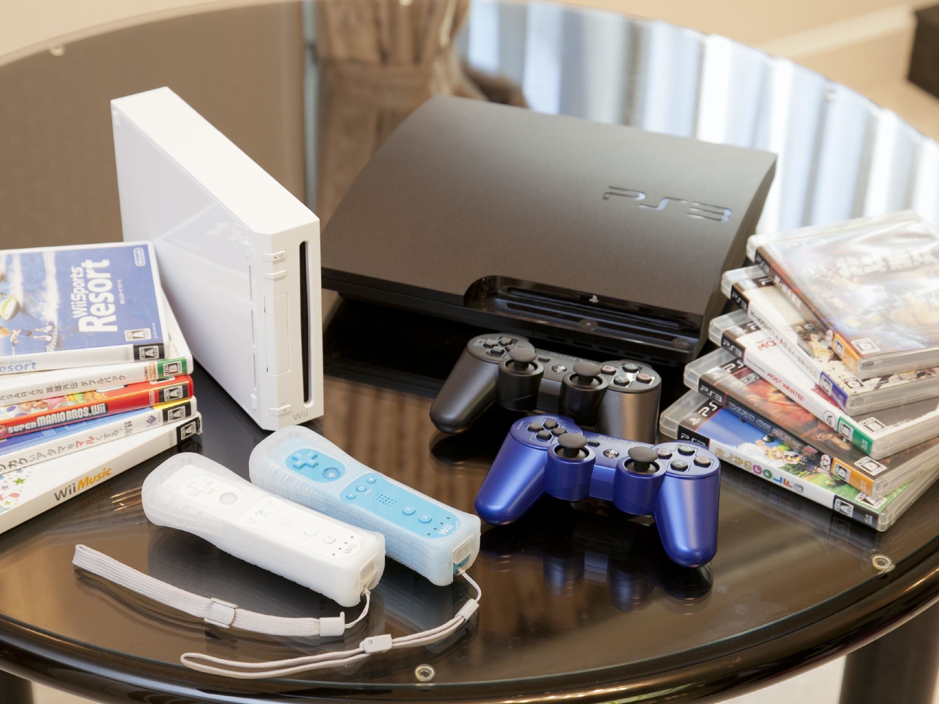 A table with a PlayStation 3 console, two game controllers, a Wii U console, and various video games.