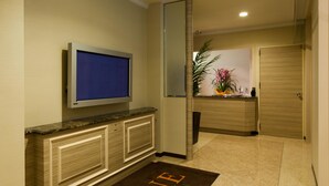 A hotel lobby with a flat-screen TV mounted on a wall, a reception desk with a plant, and a door to a room.