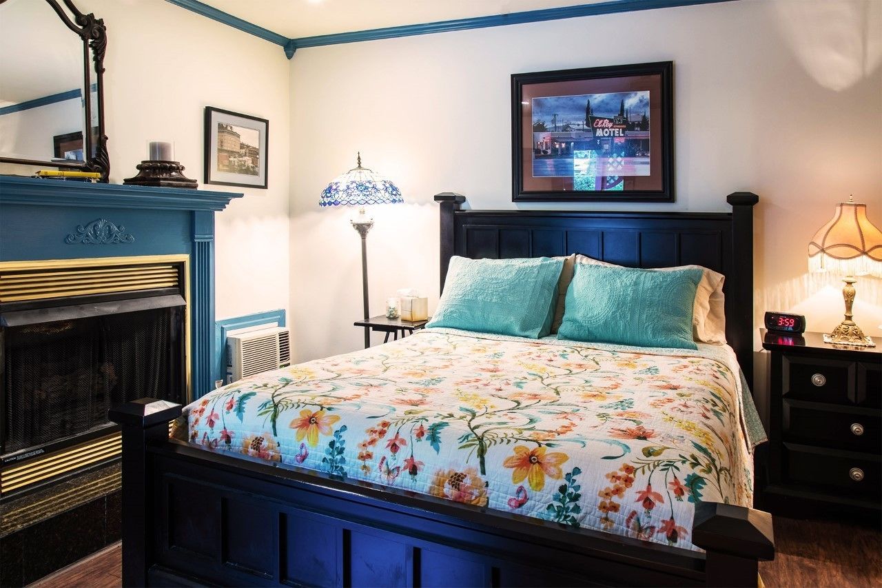 A bedroom with a floral bedspread, a fireplace, a framed picture on the wall, and a nightstand with a lamp.