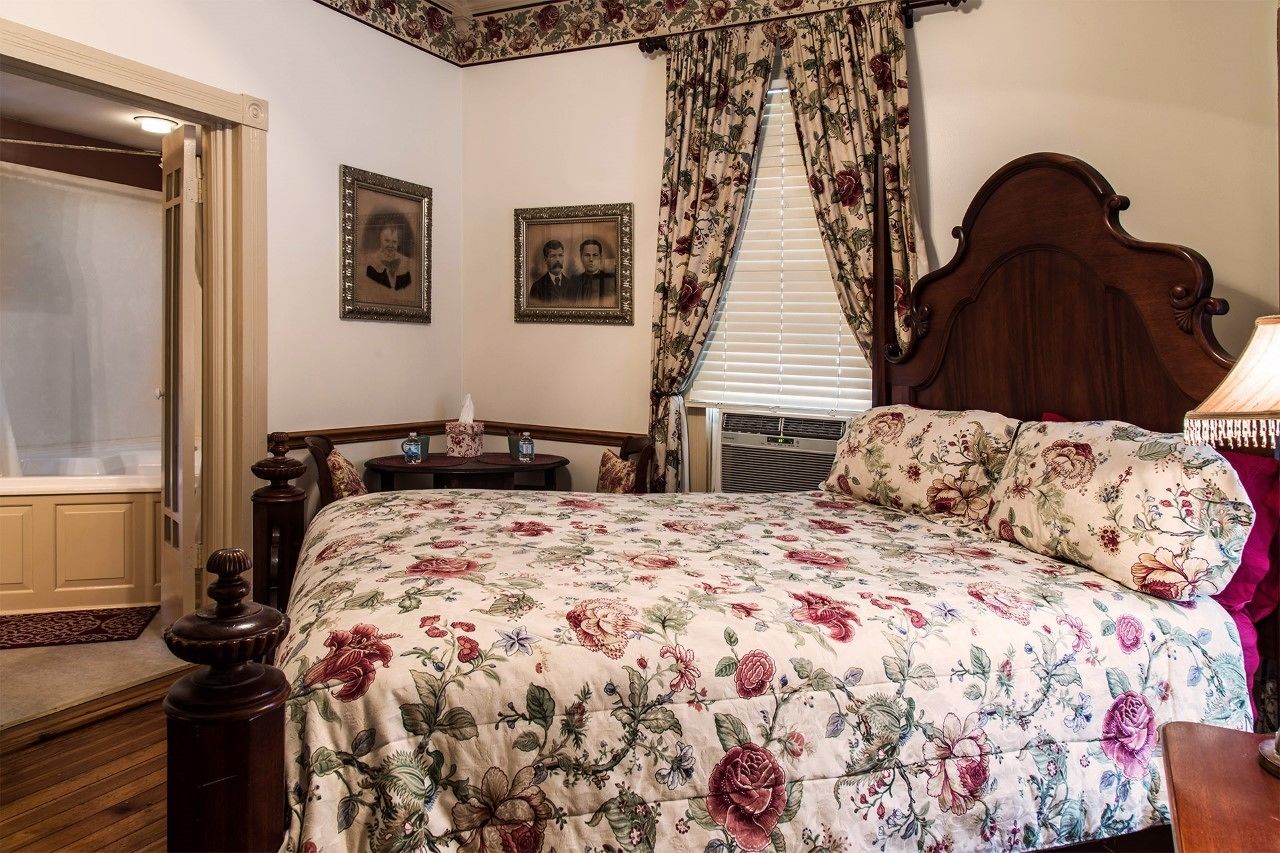 Une chambre à coucher avec une couvre-lit à motifs floraux, une tête de lit en bois, une table de chevet avec une lampe et deux tableaux encadrés accrochés au mur.