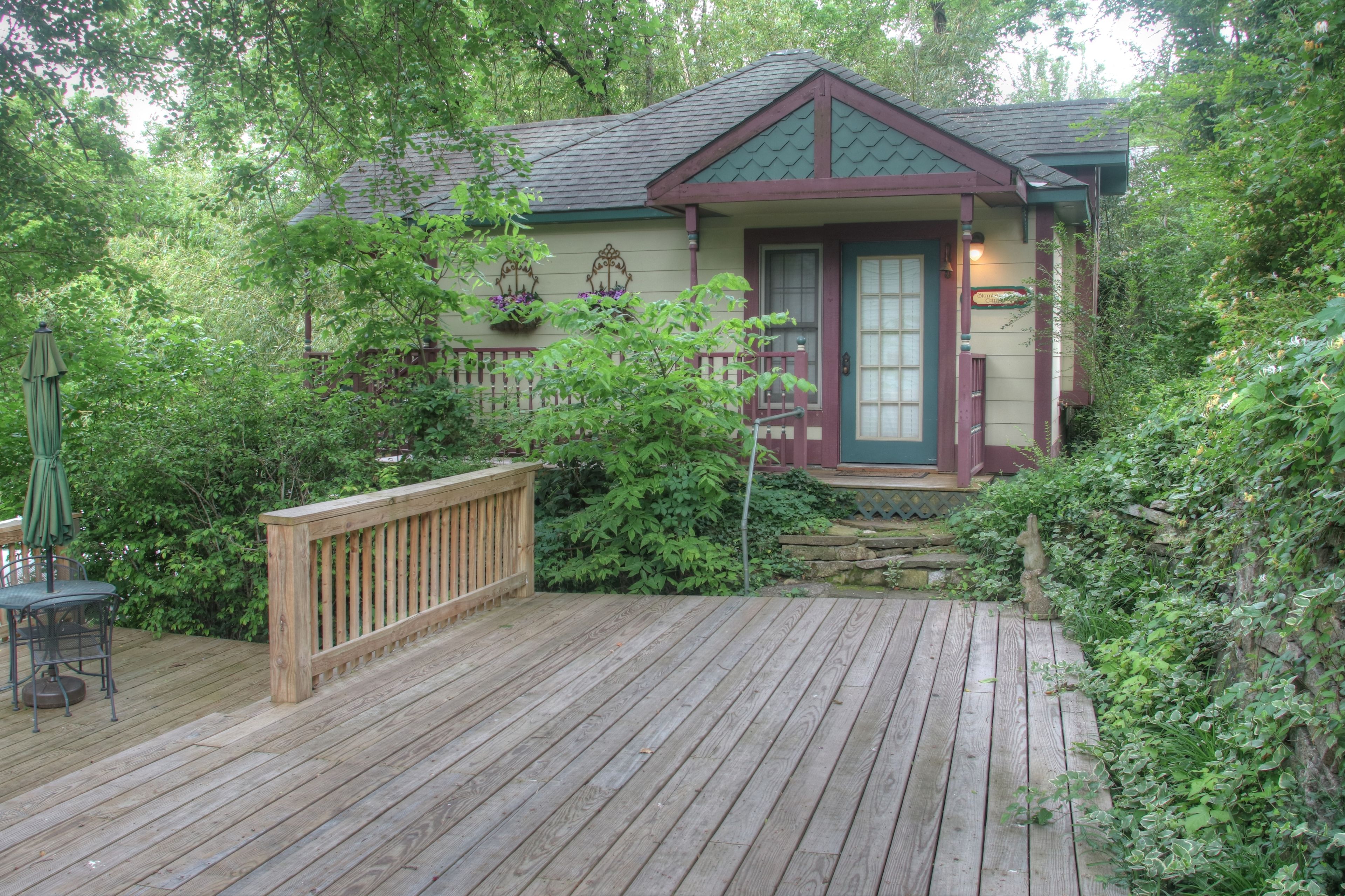 A wooden deck leading to a house with a green roof, surrounded by lush greenery.