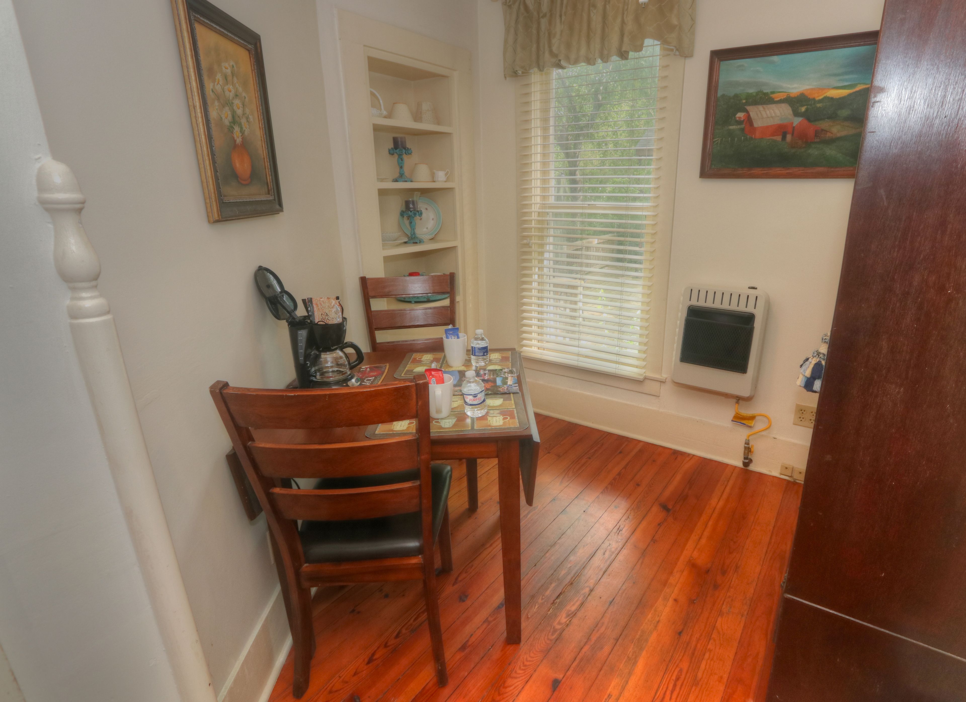 A dining area with a wooden table and chairs, a coffee maker, and a window with blinds.
