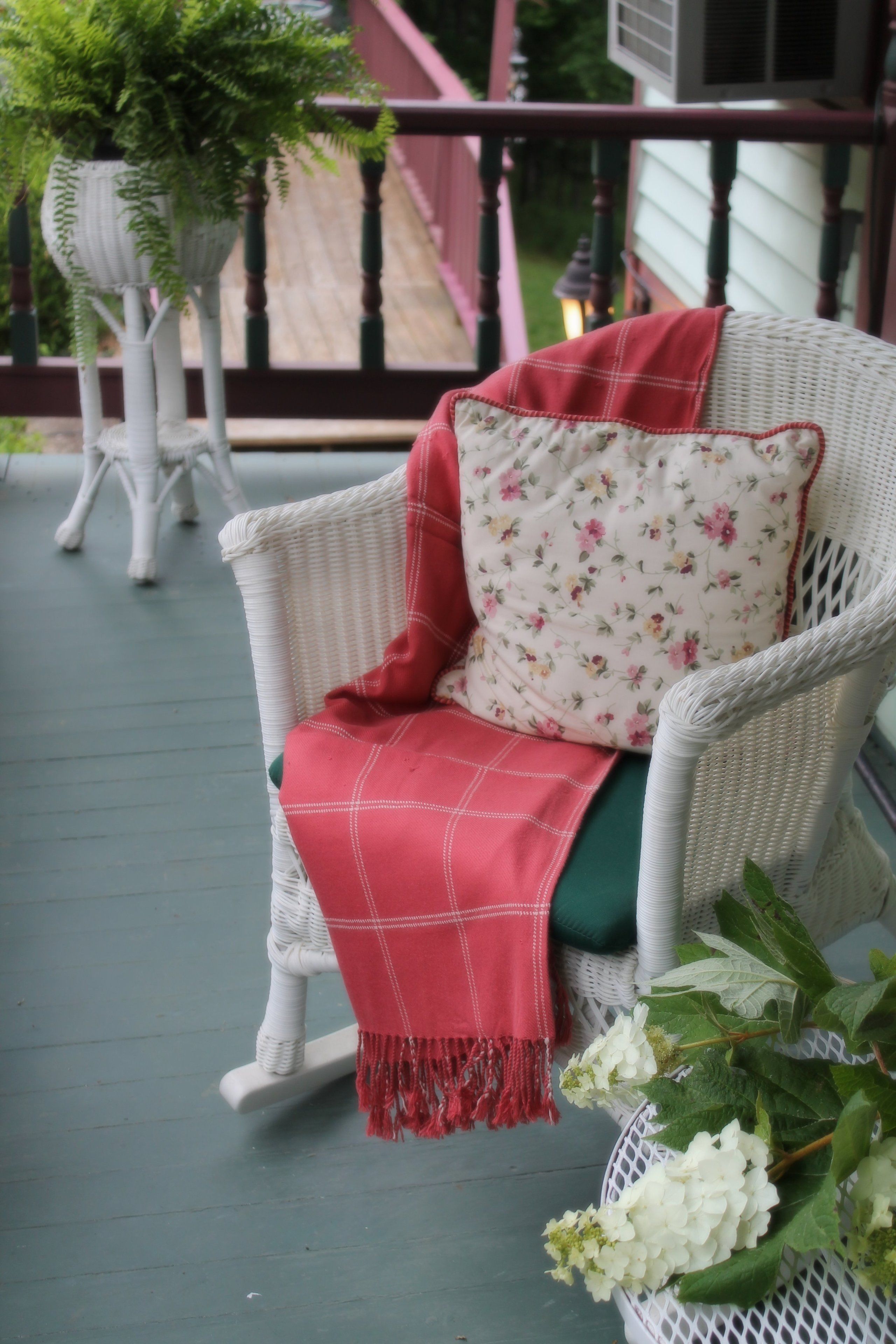 Un fauteuil à bascule en osier, agrémenté d’un coussin à motifs floraux et d’une couverture rouge, est placé sur une véranda.