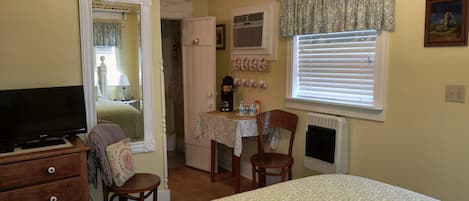 A compact room with a bed, a desk with a chair, a television, a mirror, a radiator, and a window with blinds.