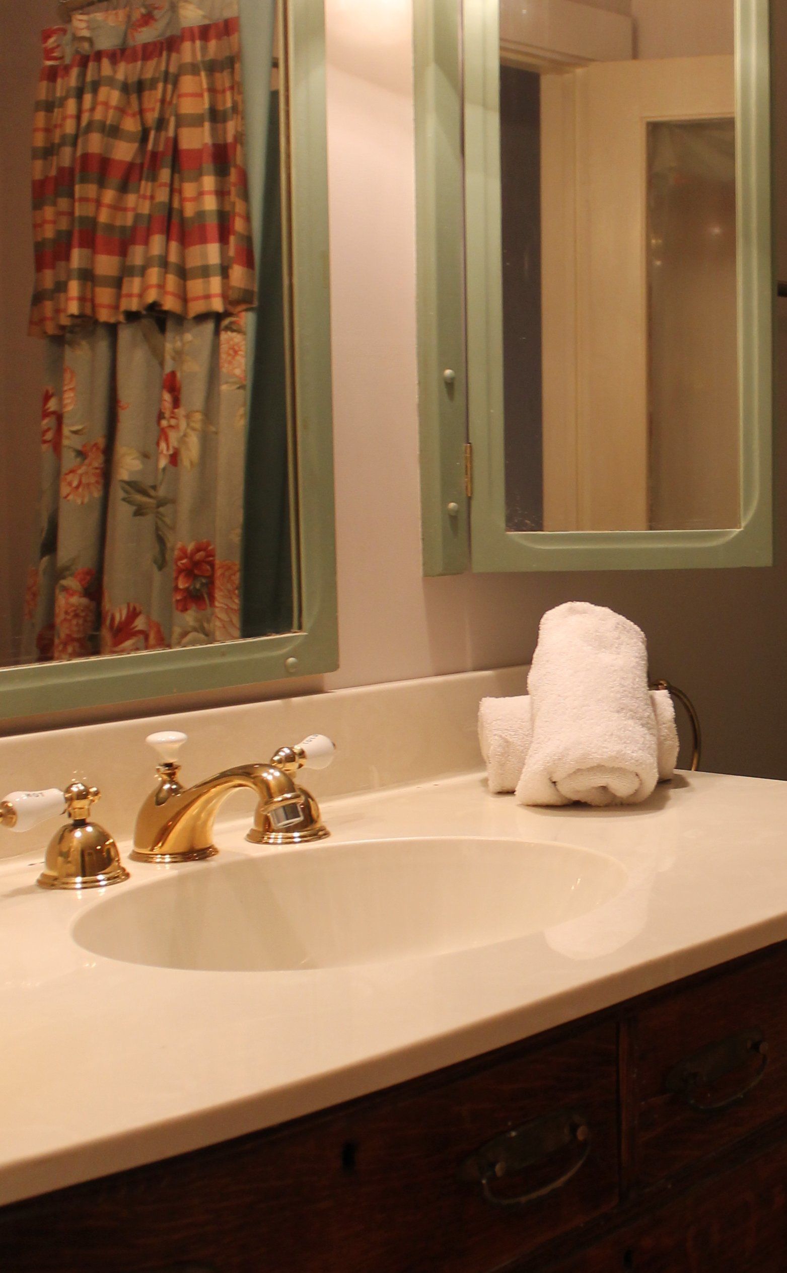 A bathroom with a sink, two gold faucets, a white towel, and a mirror reflecting a window with curtains.