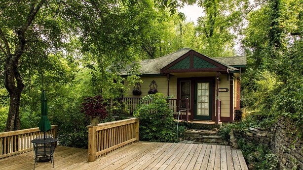 A wooden deck with a small table and chair, surrounded by lush greenery and a house with a green roof. - Hidden Springs Bed & Breakfast (Eureka Springs)