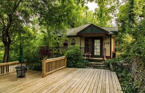 A wooden deck with a small table and chair, surrounded by lush greenery and a house with a green roof.