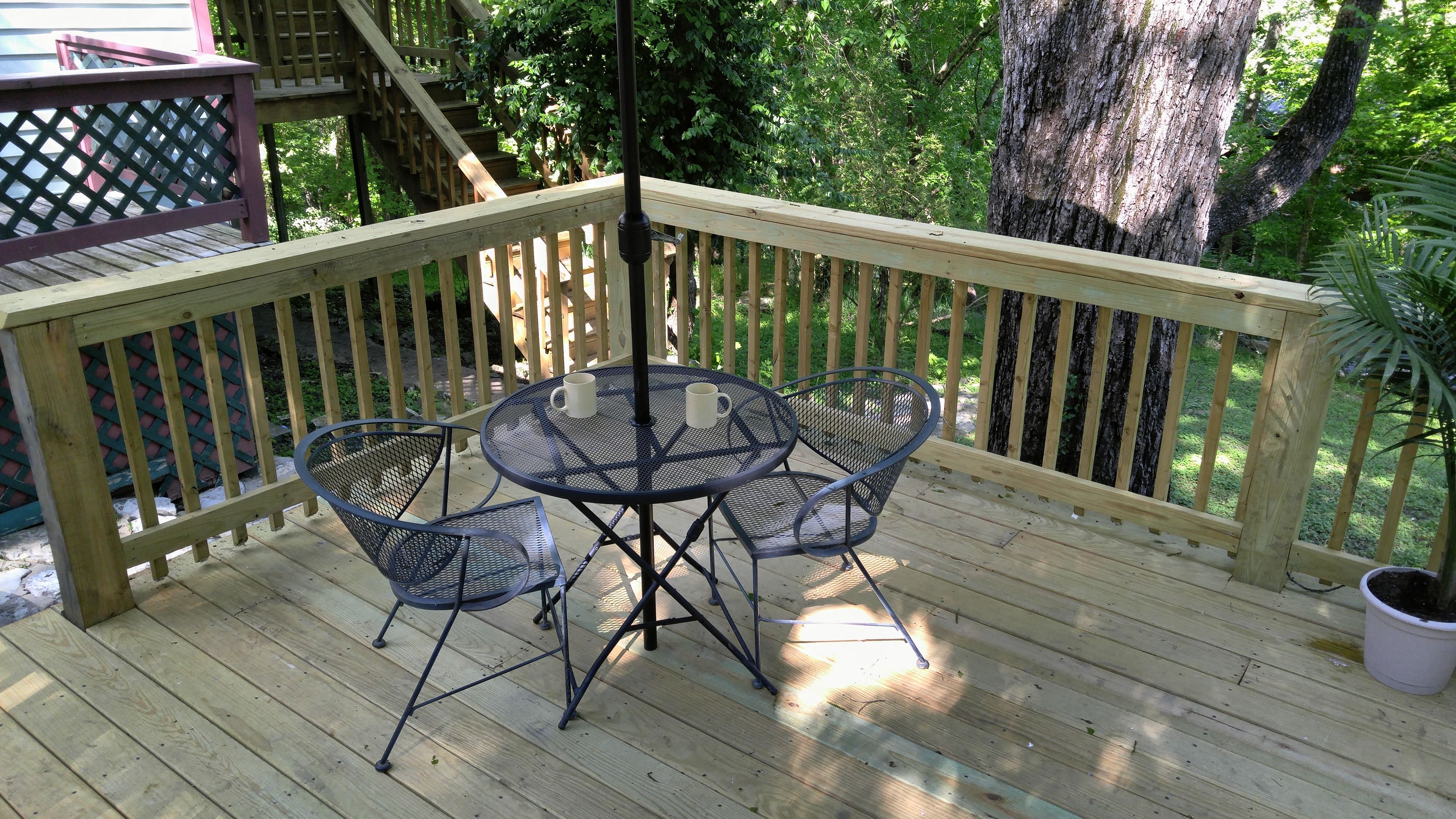 A wooden deck with a metal table and chairs, surrounded by a wooden railing and a view of trees.