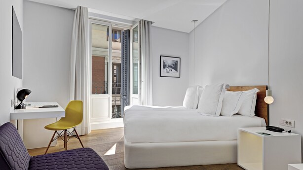 A modern hotel room with a large bed, a desk with a chair, a small sofa, and a nightstand with a lamp. - One Shot Prado (Madrid)