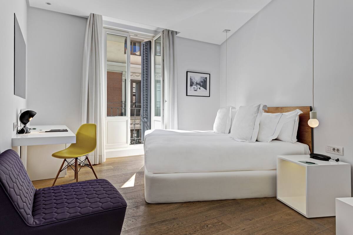 A modern hotel room with a large bed, a desk with a chair, a small sofa, and a nightstand with a lamp.