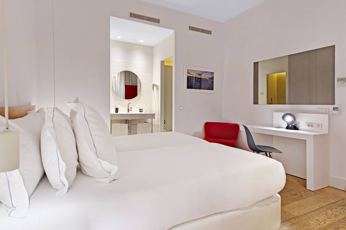 A hotel room with a large bed, a desk with a chair, a mirror, and a red chair.