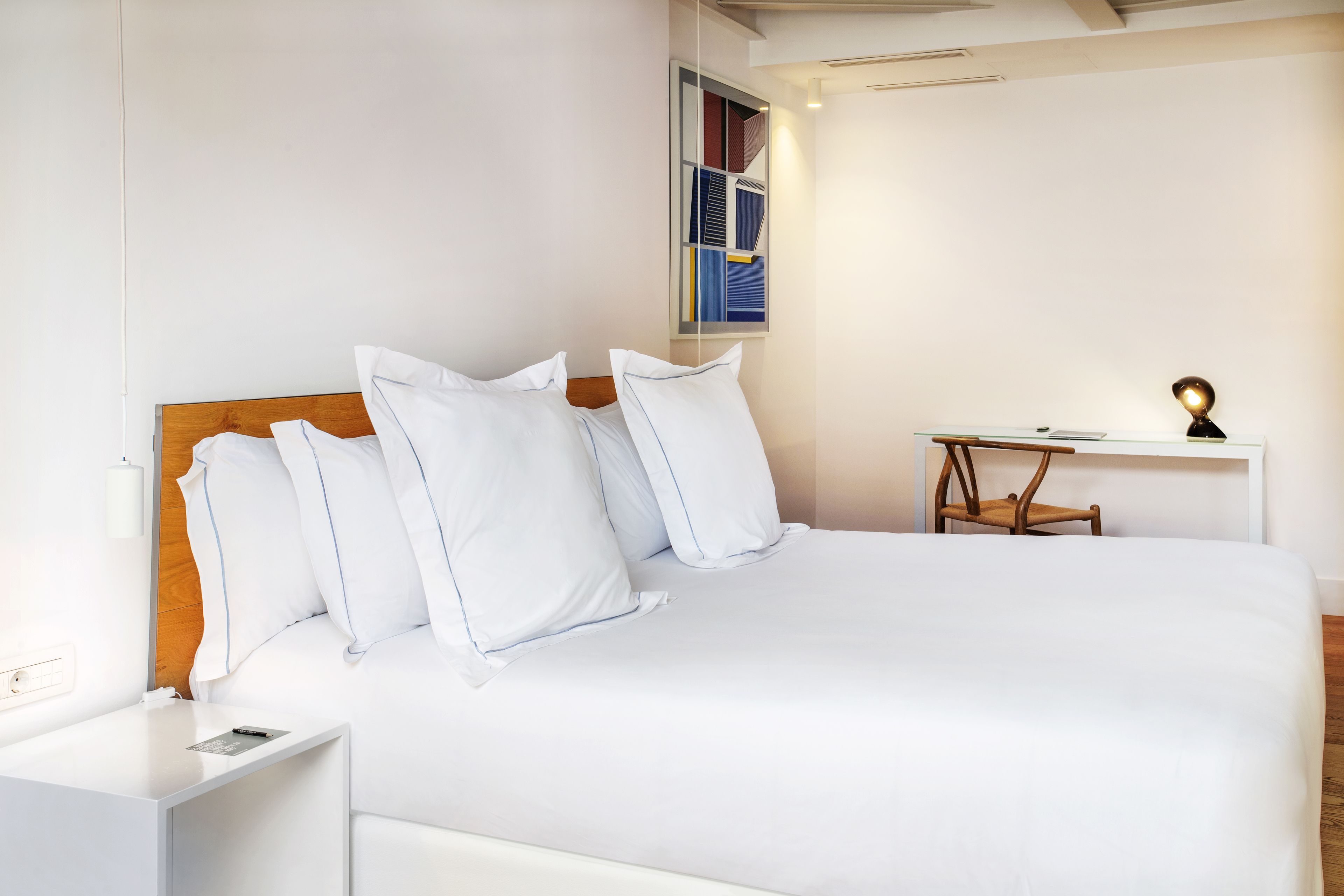 Una cama bien hecha con sábanas y almohadas blancas, una mesita de noche con una lámpara, y una pequeña silla de madera.