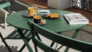 A green table with a glass of coffee, a plate with a pastry, and a magazine on a balcony with a white building and metal railings in the background. - One Shot Prado (Madrid)