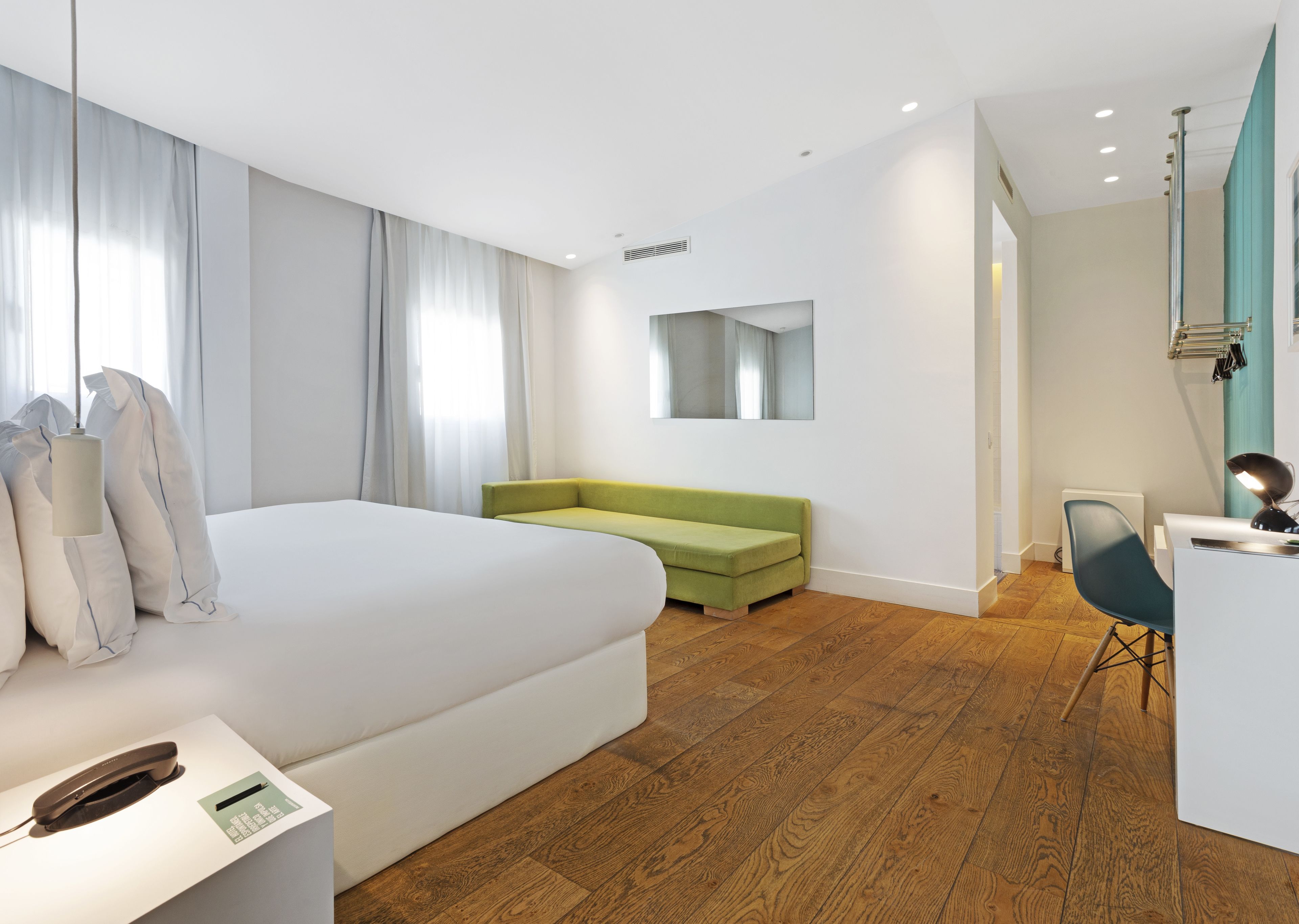 A modern hotel room with a large bed, a green sofa, a desk with a chair, and a mirror on the wall.