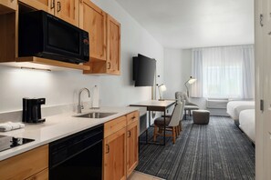 A compact hotel room with a kitchenette, a flat-screen TV, a desk with a chair, a bed, and a window with curtains. - TownePlace Suites by Marriott Minot (Minot)
