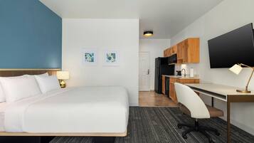 A hotel room with a bed, desk, chair, kitchenette, and a TV.
