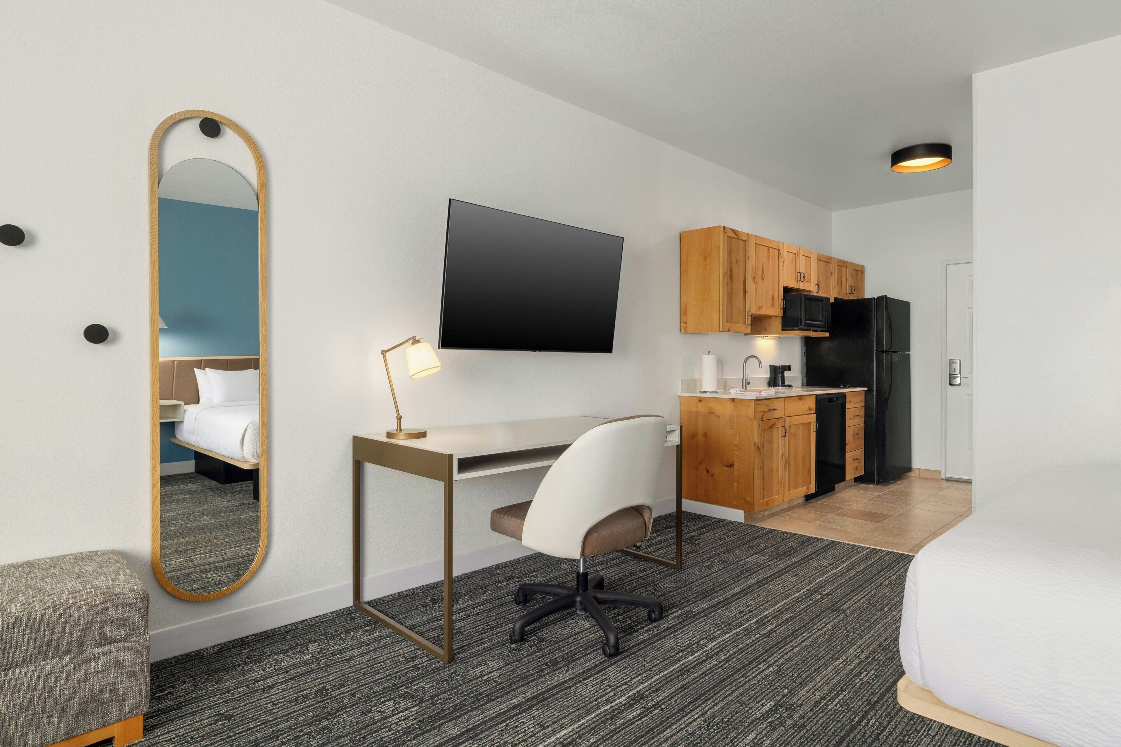 Une chambre d’hôtel comprenant un lit, un bureau avec une chaise, une télévision, un miroir, une kitchenette et un sol recouvert de moquette.