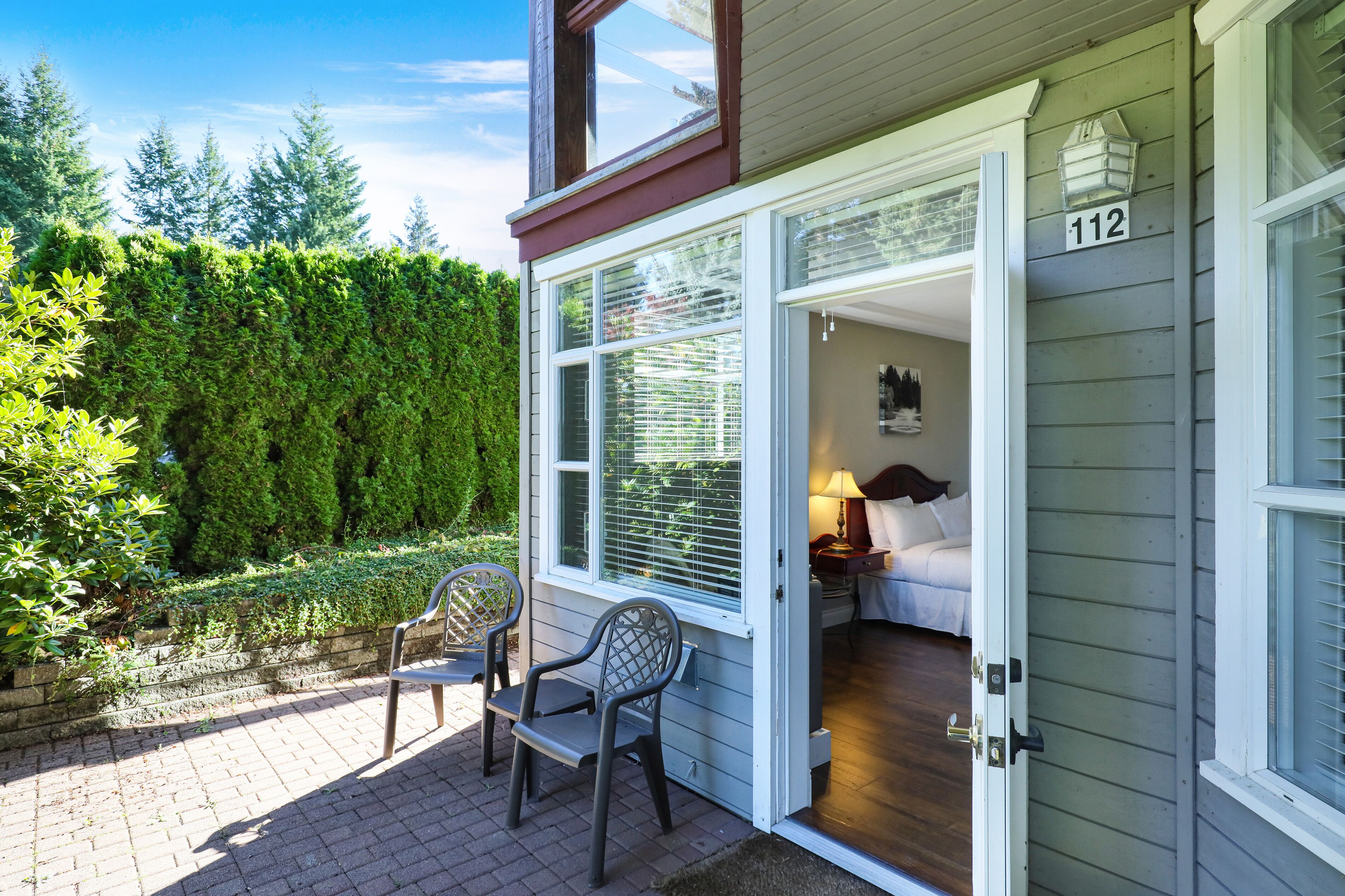 Une maison portant le numéro 112, une terrasse en bois avec deux chaises, et un jardin agrémenté d’une haie.