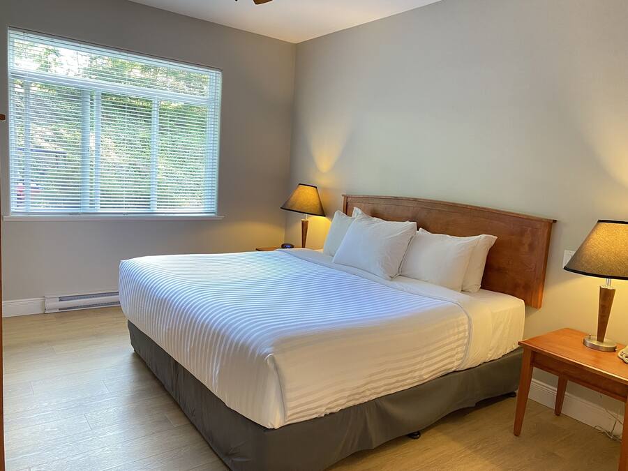 Una cama bien hecha con cabecera de madera, una mesita de noche con lámpara y una ventana con persianas.