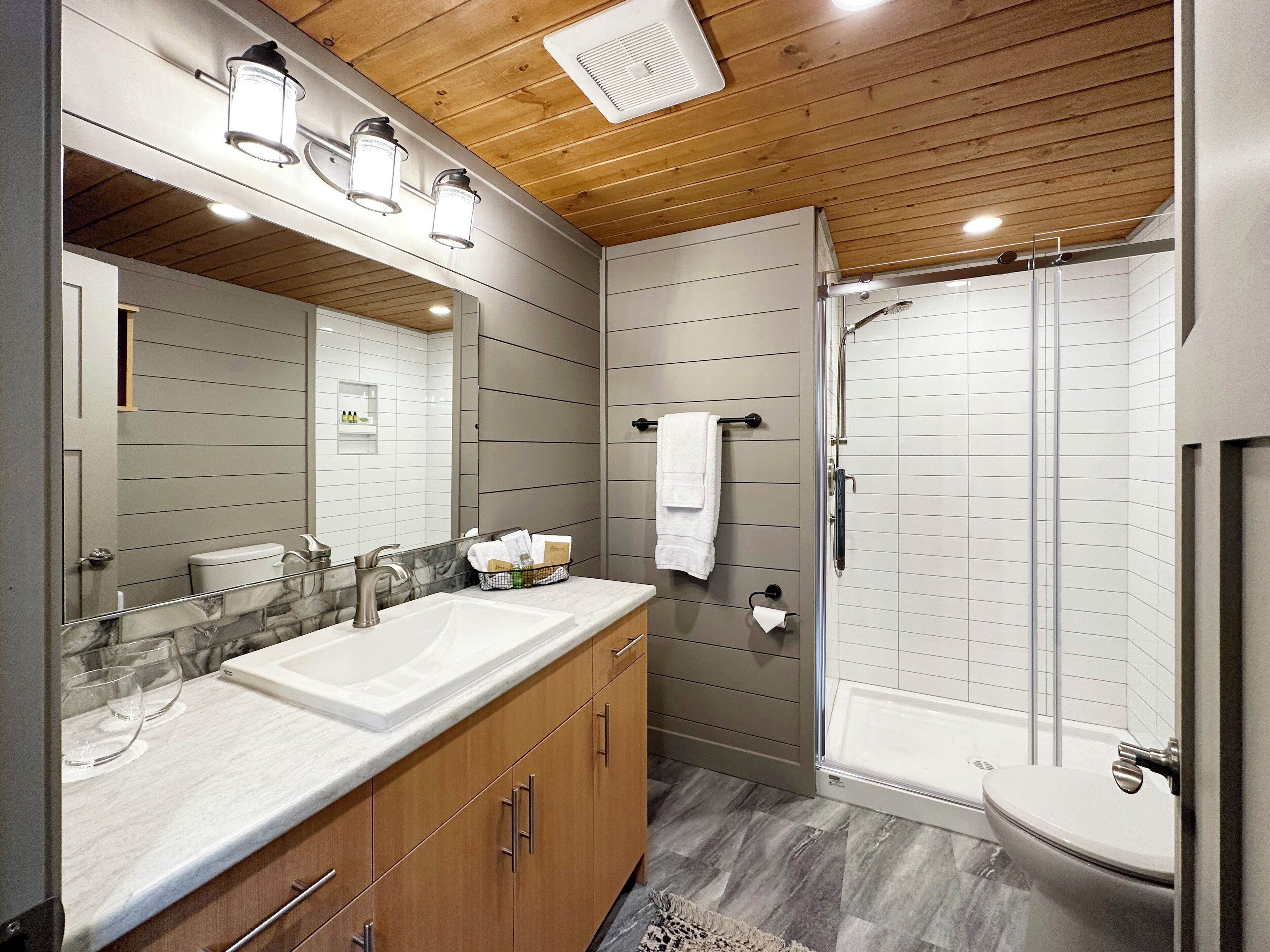 Un baño moderno con techo de madera, un espejo grande, tocador doble, cabina de ducha y un inodoro.