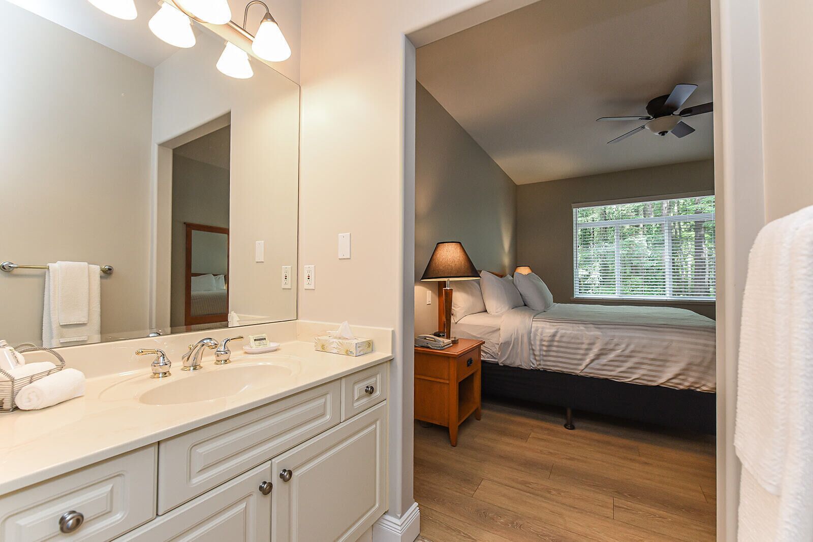 A hotel room with a large mirror, a double sink, a bedside table with a lamp, a bed, and a ceiling fan.