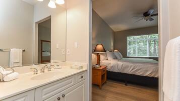 A hotel room with a large mirror, a double sink, a bedside table with a lamp, a bed, and a ceiling fan.