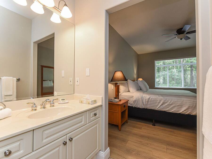 A hotel room with a large mirror, a double sink, a bedside table with a lamp, a bed, and a ceiling fan.