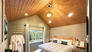 Une chambre avec un plafond en pente, un grand lit recouvert d’une couverture à carreaux, deux tables de chevet avec des lampes, une table de chevet avec une lampe, une fenêtre avec des rideaux et un banc avec un magazine.