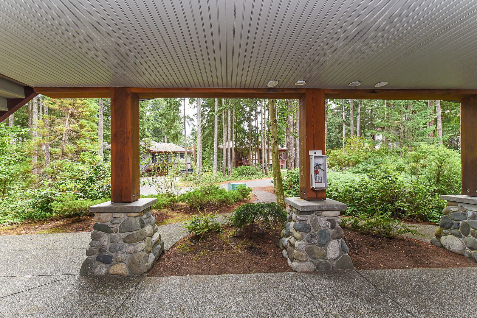 A covered outdoor area with stone pillars, a wooden ceiling, and a view of a forested area with a building in the background.