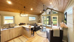 Une cuisine chaleureuse avec des meubles en bois, un coin repas comprenant une table et des chaises, et de grandes fenêtres offrant une vue sur l’extérieur.
