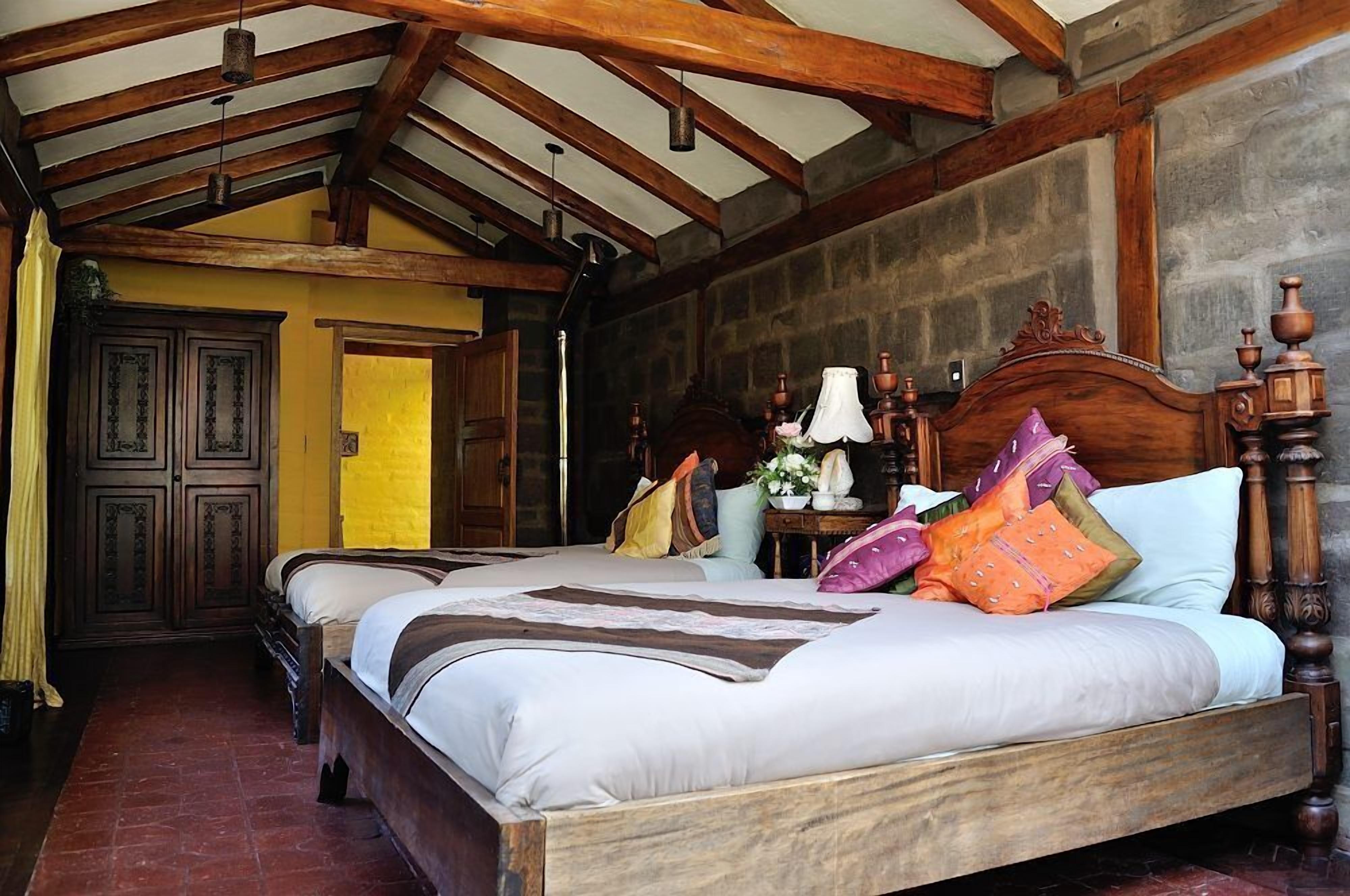 Una cama de madera tradicional con cabecera, una mesita de noche con lámpara, y una pared con textura de piedra.