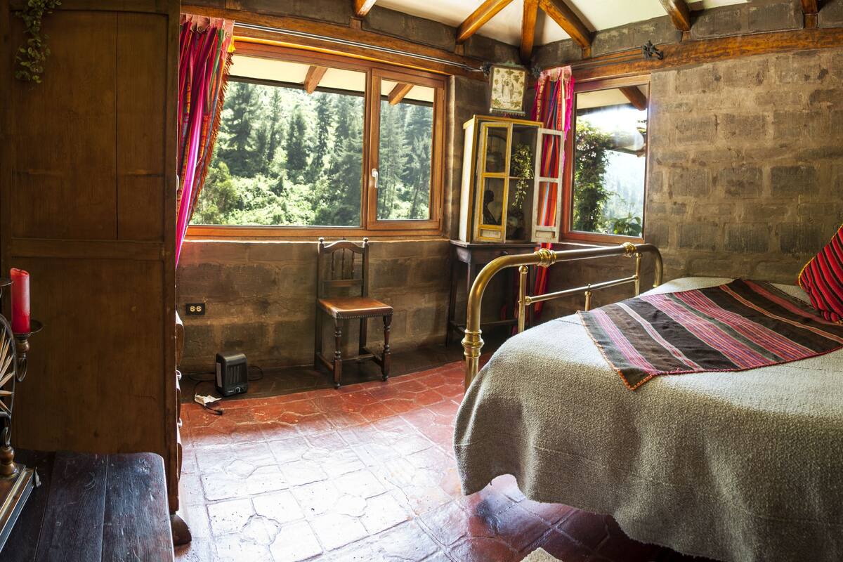 A rustic bedroom with a wooden bed, a small wooden chair, a window with curtains, and a stone wall.