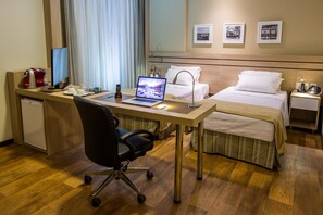 Ein Hotelzimmer mit Schreibtisch aus Holz, einem Laptop, einem schwarzen Bürostuhl, einem Bett mit weißen Bettwäsche, einem Nachttisch mit Lampe und zwei gerahmten Bildern an der Wand.