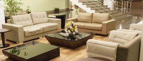 A modern hotel lobby with beige sofas, a glass coffee table, and a staircase leading to the upper floor.