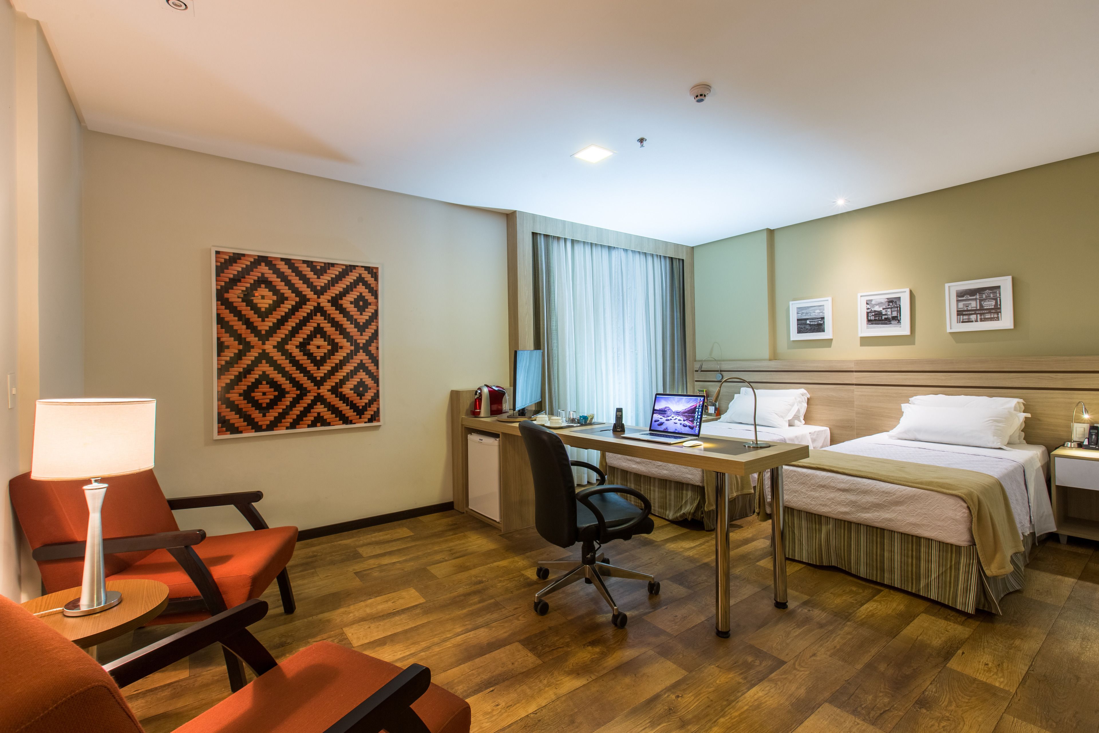 A hotel room with a desk, two beds, a chair, a lamp, a laptop, and a window with curtains.