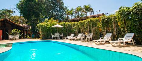 A clear swimming pool with lounge chairs and umbrellas, surrounded by lush greenery and a building with a tiled roof.