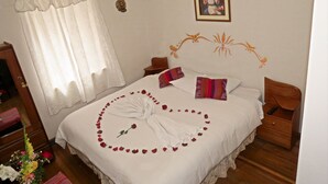 A neatly made bed with rose petals arranged in a heart shape, a framed religious picture on the wall, and a wooden nightstand with a remote control. - Casa La Sambleña INN (Cusco)