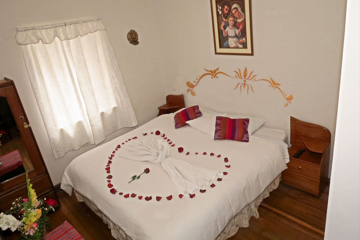 A neatly made bed with rose petals arranged in a heart shape, a framed religious picture on the wall, and a wooden nightstand with a remote control.