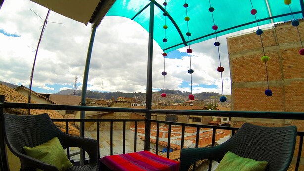 A balcony with wicker chairs, a small table with a colorful tablecloth, and a view of a cityscape with brick buildings and distant hills. - Casa La Sambleña INN (Cusco)