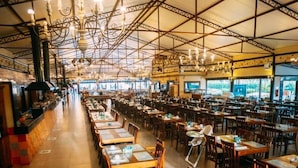A spacious restaurant with high ceilings, exposed beams, and a long counter along one side. Tables are neatly arranged with menus and cutlery.