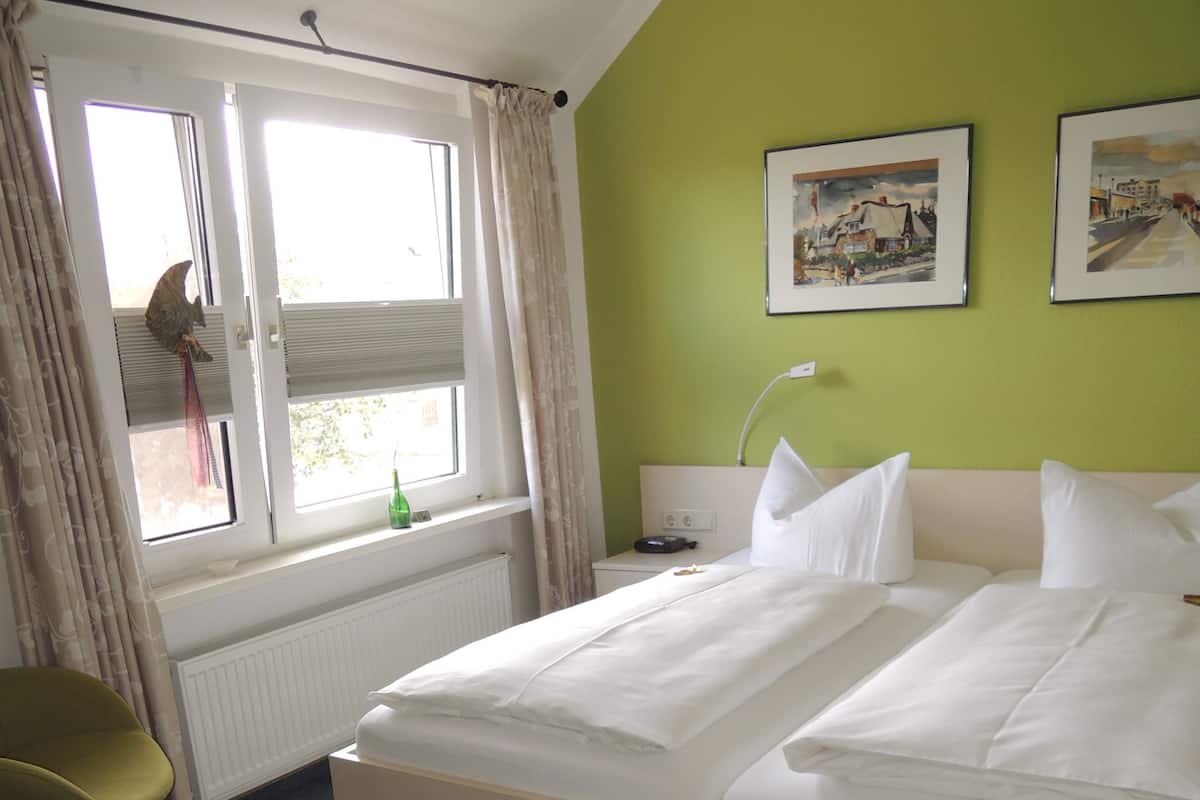 A hotel room with a bed, a chair, a window with curtains, a radiator, and two framed pictures on the wall.