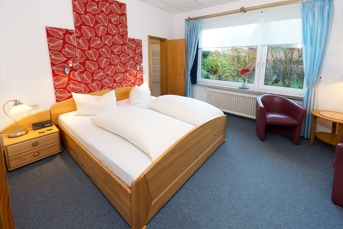 A hotel room with a wooden bed, a red floral wall decoration, a window with a view of greenery, and a red chair.