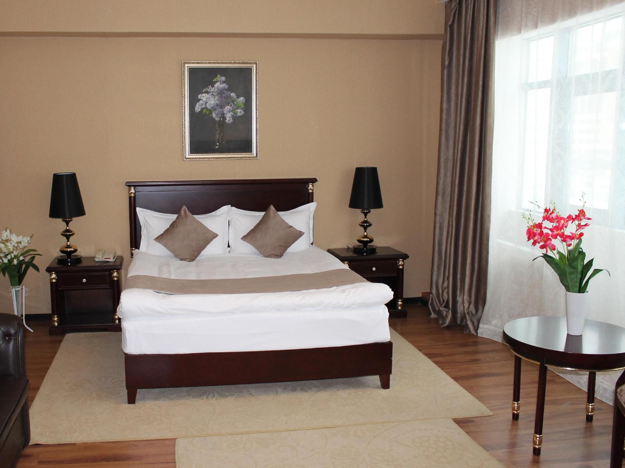 Un letto rifatto con un copriletto bianco e due cuscini, affiancato da comodini con lampade, in una stanza con un'ampia finestra e un quadro appeso al muro.