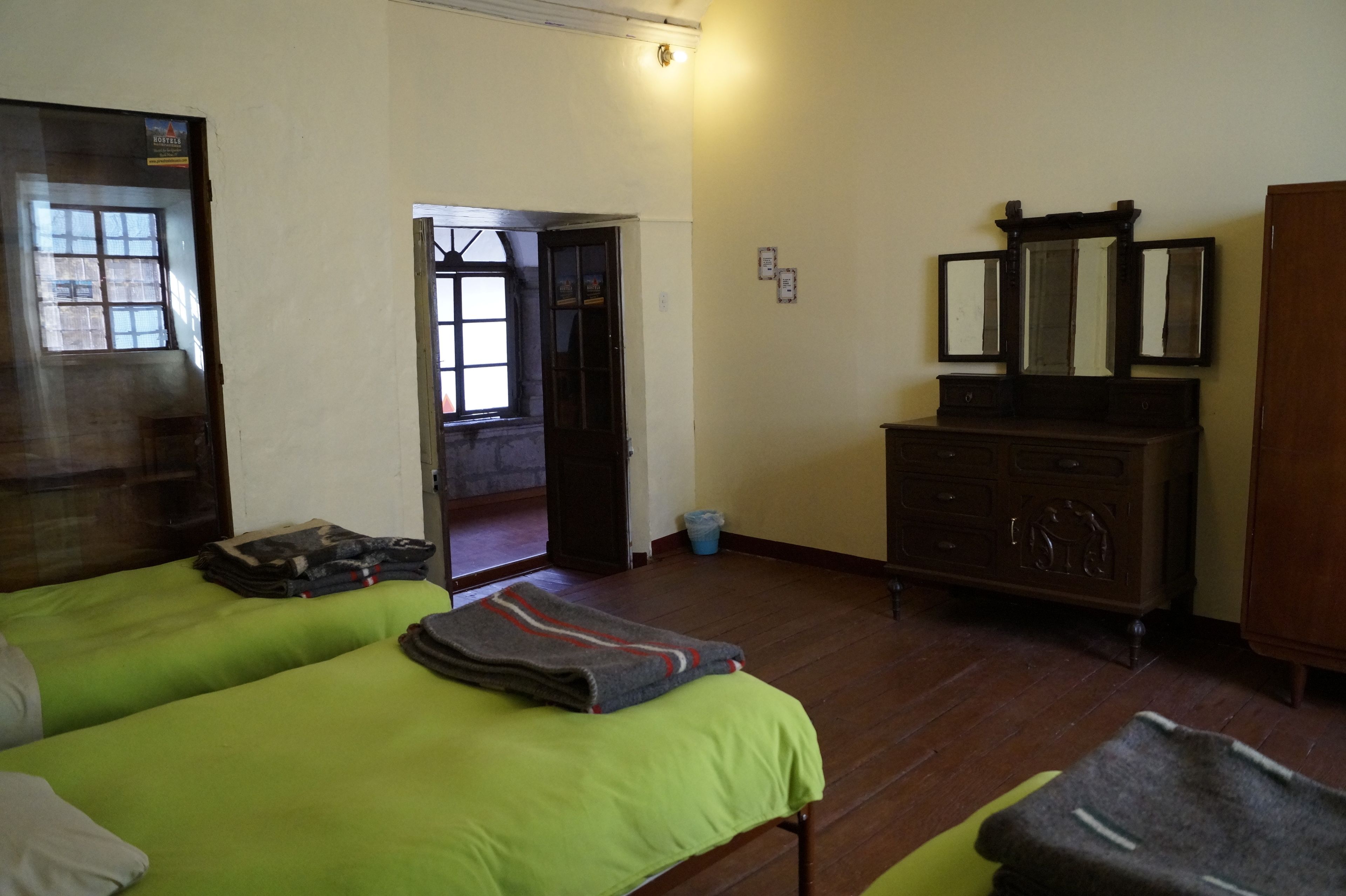A room with two green beds, a wooden dresser with a mirror, and a door leading to another room.