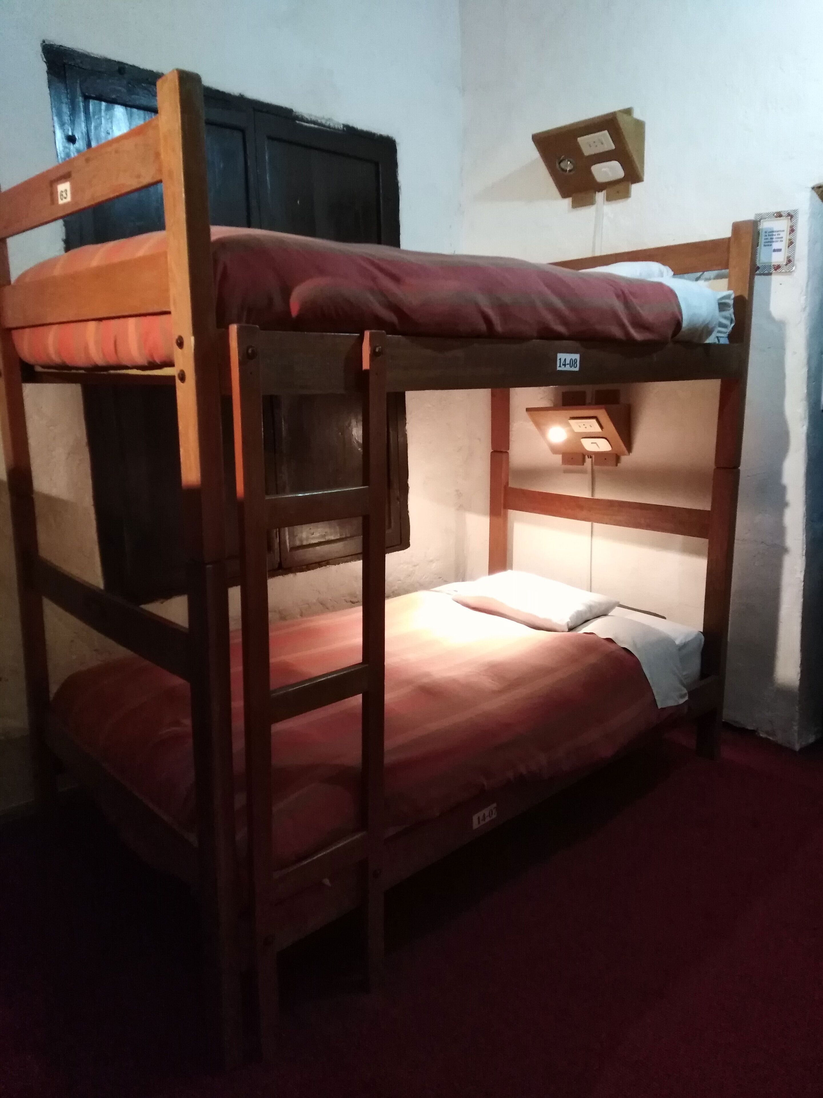 A bunk bed with a wooden frame, a mattress, and a pillow. There is a wall socket and a light switch on the wall.