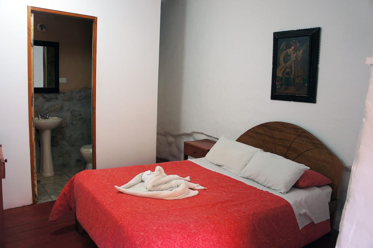Ein Hotelzimmer mit Bett, Nachttisch, Holz-Kopfteil, einem Bild an der Wand und einem sichtbaren Badezimmer hinter einer offenen Tür.