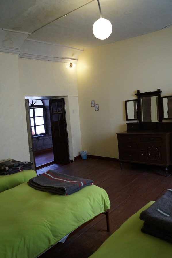 A room with two green beds, a wooden dresser with a mirror, a light fixture, and a door leading to another room.