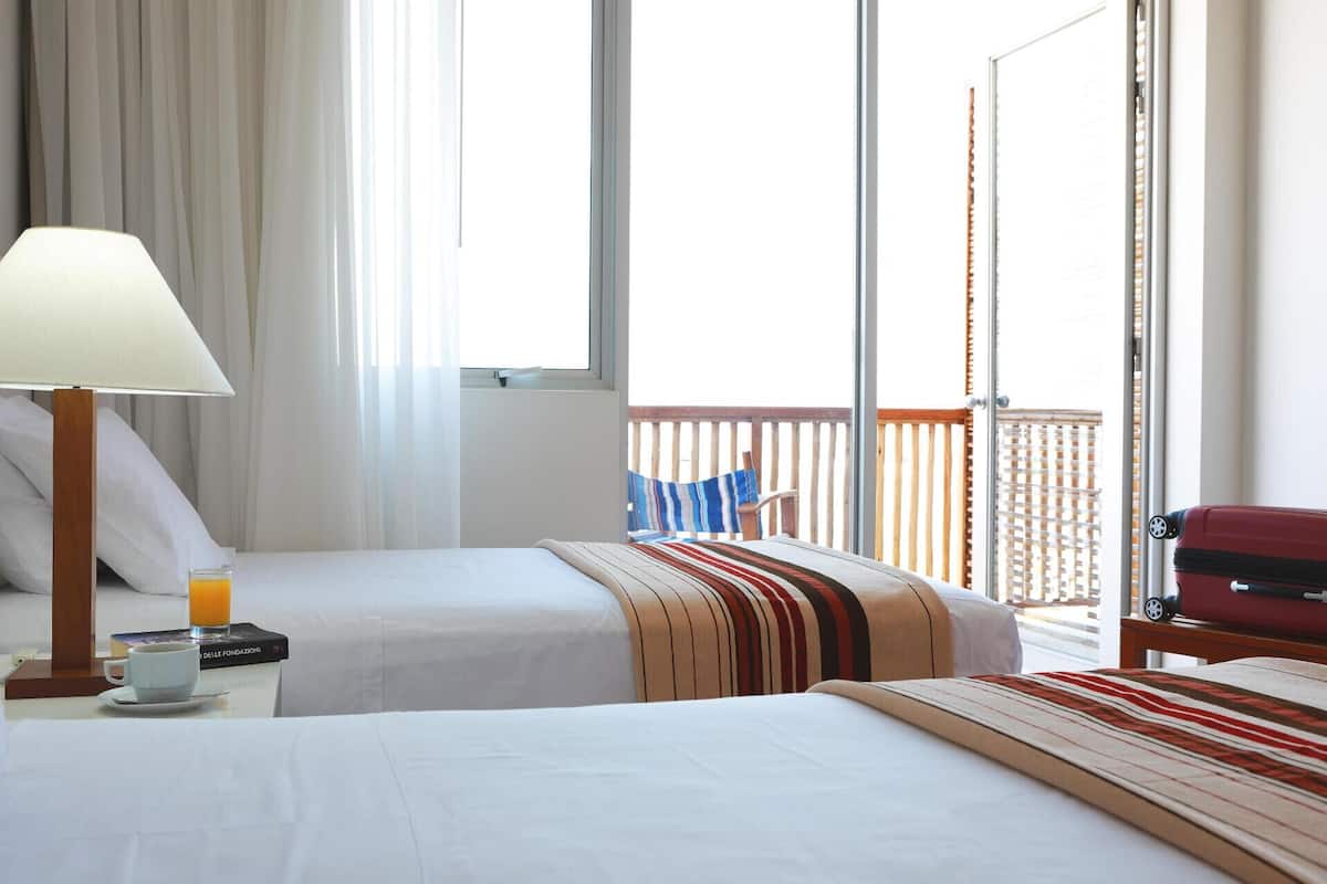 A hotel room with two beds, a nightstand with a lamp, a chair, and a view of the outside balcony.